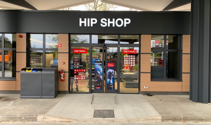 Na slici je ulaz u Hip Shop na Petrol prodajnom mjestu. Iznad staklenog ulaza nalazi se veliki natpis „HIP SHOP“. Pored ulaza nalaze se kante za odvajanje otpada i protivpožarni aparat. Na ulaznim vratima i staklenim površinama vidljive su različite informativne i promotivne naljepnice.