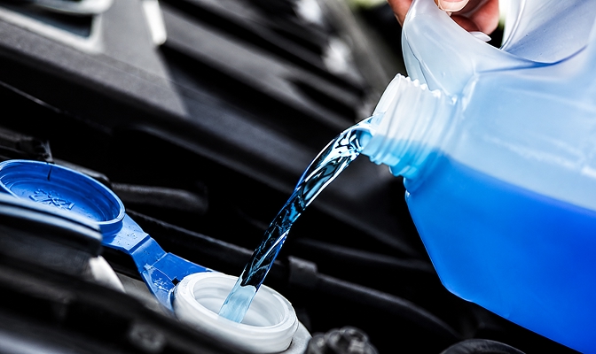 Na slici je plava tečnost koja se sipa iz plastične posude u rezervoar u motornom prostoru vozila. U prvom planu vidljivi su otvor rezervoara i poklopac, dok se u pozadini nalaze djelovi motora automobila.